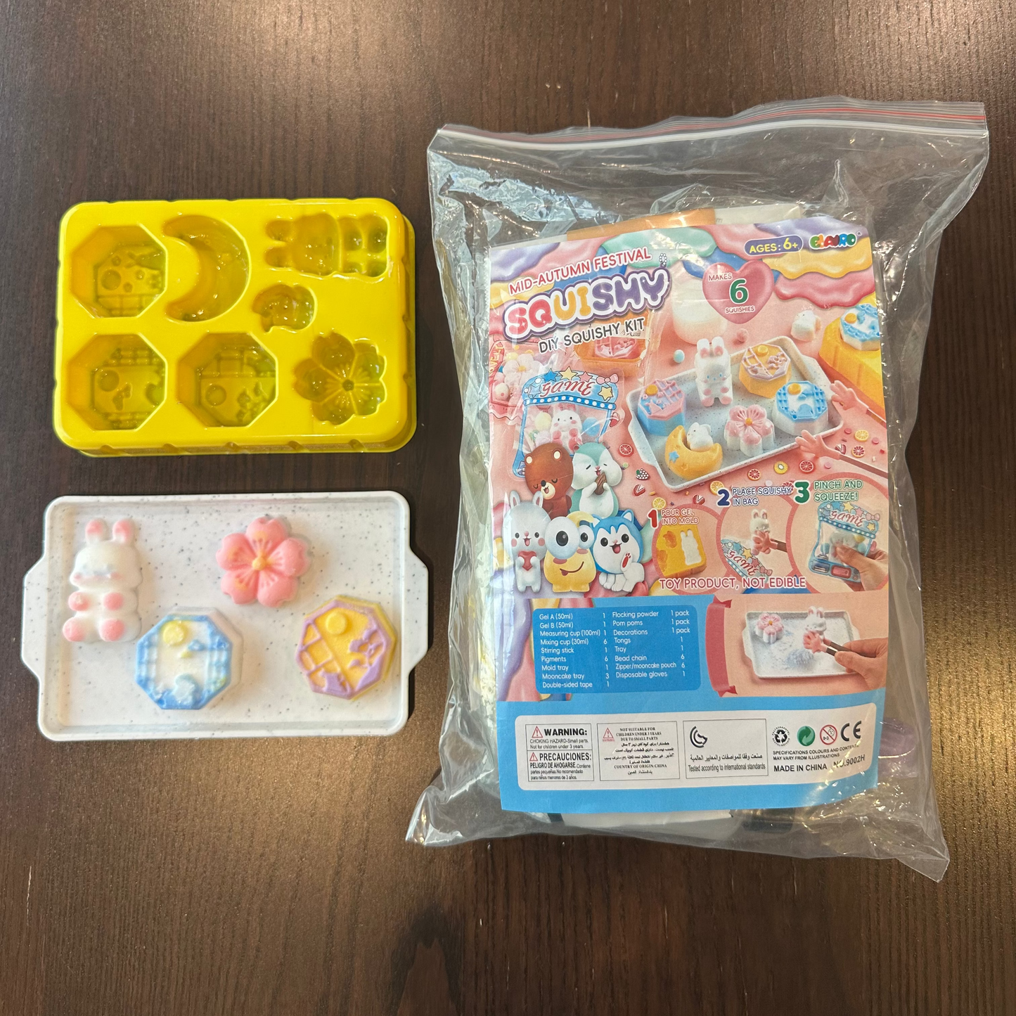 A toy craft set laid out on a dark wood surface, including a yellow silicone mold tray, a white tray with three small pastel-colored squishy charms, and the retail packaging for an EQUSKY squishy set in a clear plastic bag.
