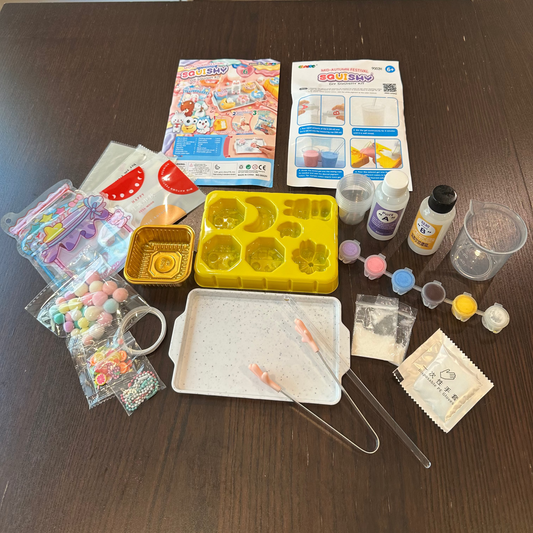 A DIY craft kit laid out on a dark wooden table, including a yellow mold tray, paint bottles, a mixing cup, tweezers, beads, and instruction sheets.