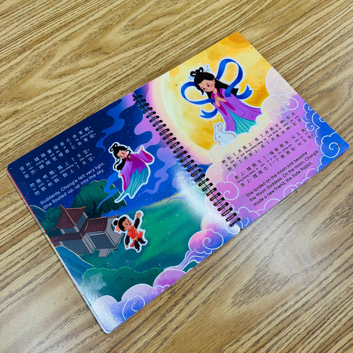 A spiral-bound children's book with colorful Chinese illustrations, placed diagonally on a light brown wooden tabletop.
