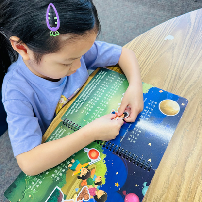 A young child wearing a light blue t-shirt and a purple hair clip sits at a wooden table, interacting with a colorful spiral-bound children's book about space.