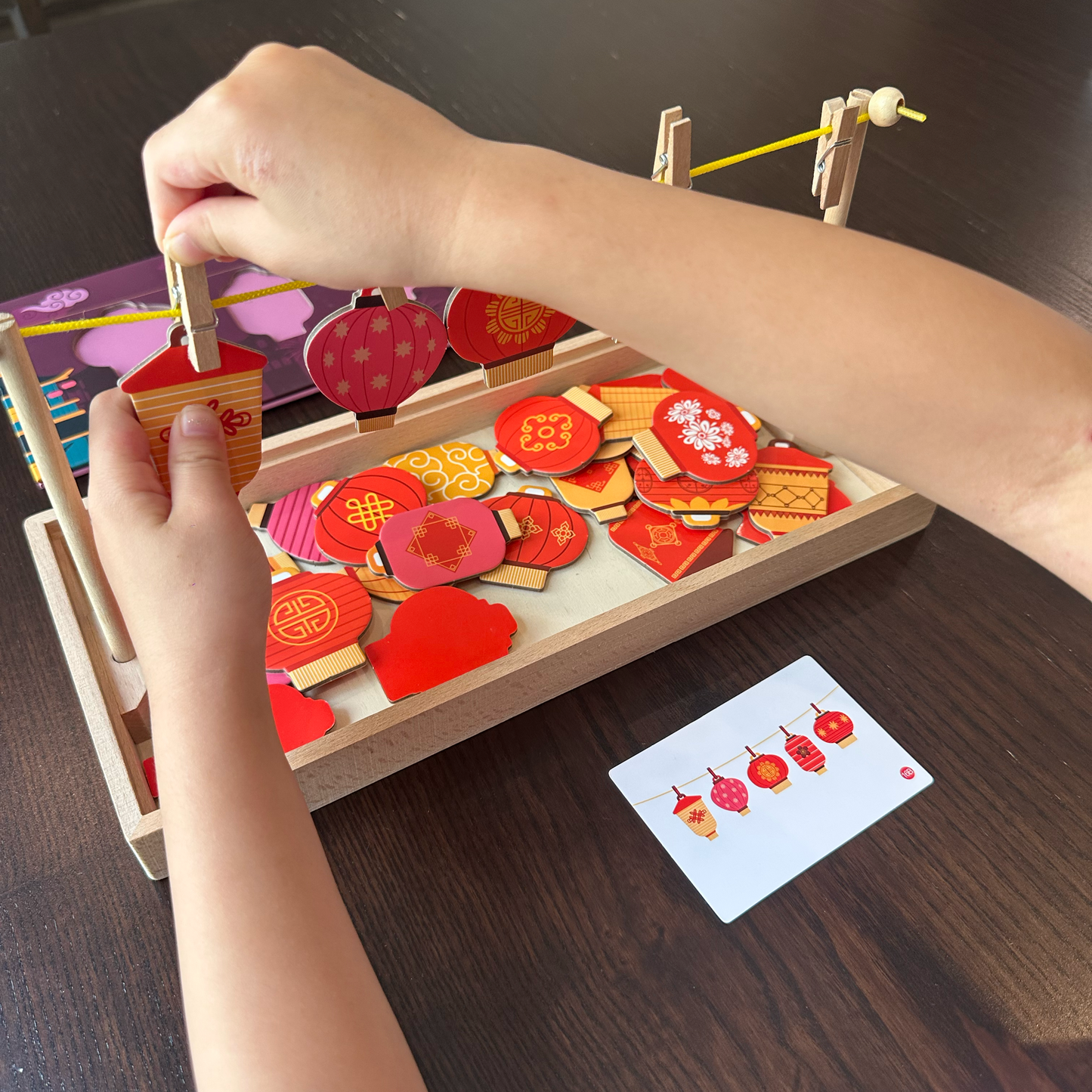 A child's hands are shown interacting with a wooden Chinese New Year-themed educational toy set on a dark wooden table. The set contains red and gold festive wooden pieces, including lanterns and decorative shapes, arranged in a tray, with a white instruction card nearby.