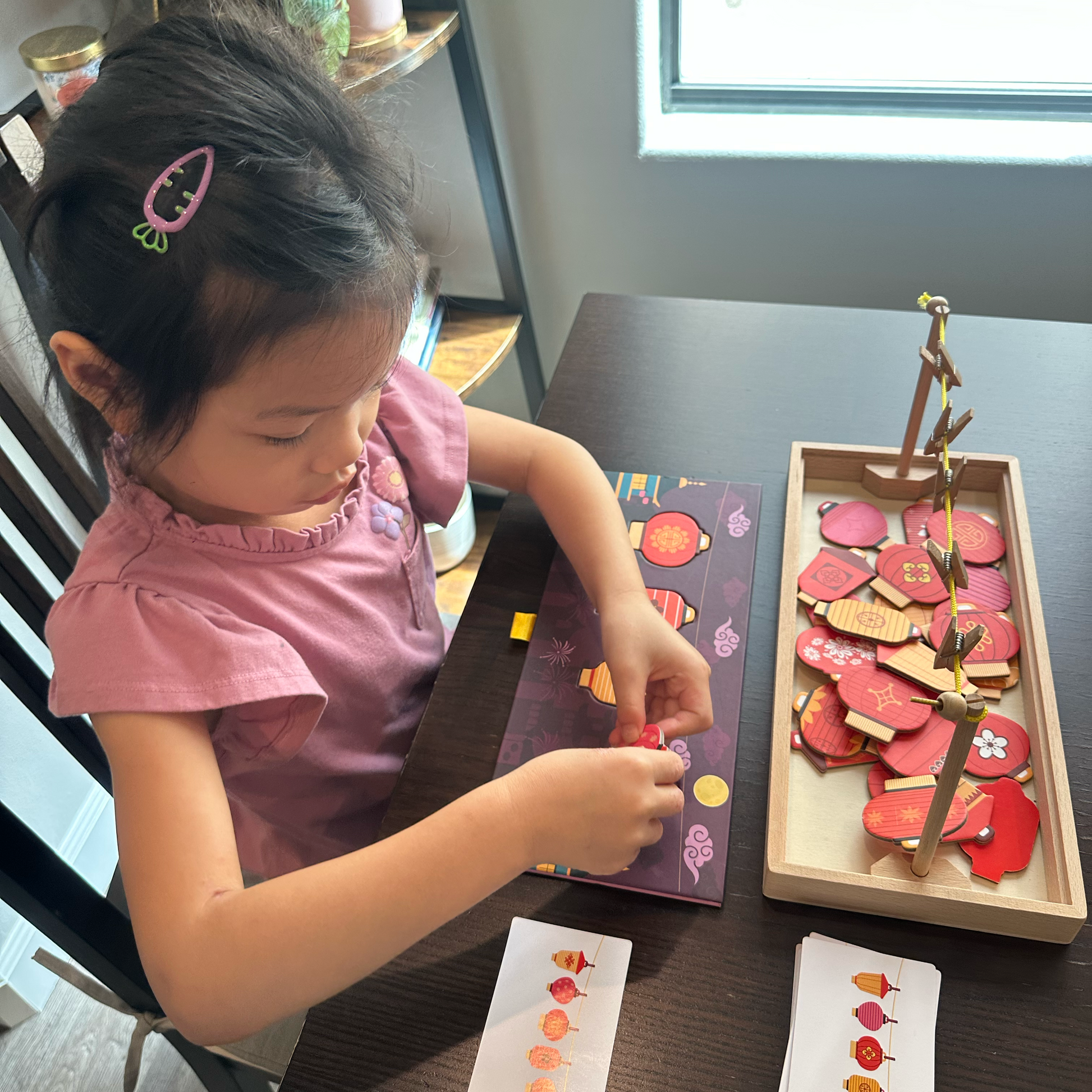 A young child wearing a pink top is lying on a dark table, playing with a colorful wooden fruit sorting and matching toy set. The toy includes a wooden tray with red and pink fruit pieces, a matching sorting board, and small cards with fruit illustrations.
