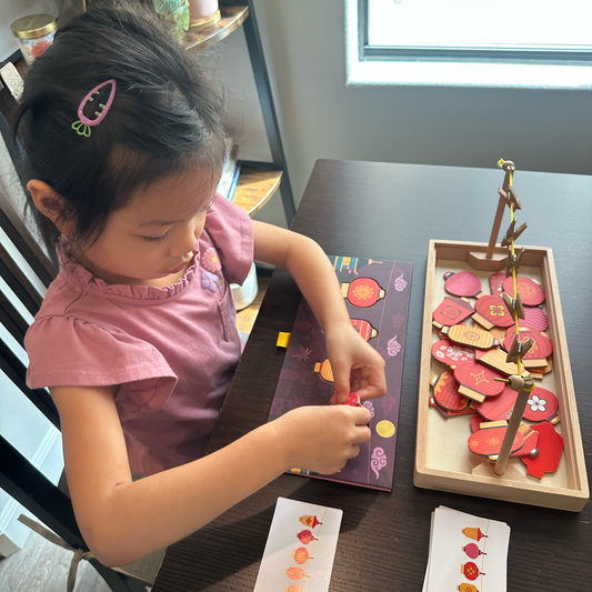 A young child wearing a pink top is lying on a dark table, playing with a colorful wooden fruit sorting and matching toy set. The toy includes a wooden tray with red and pink fruit pieces, a matching sorting board, and small cards with fruit illustrations.